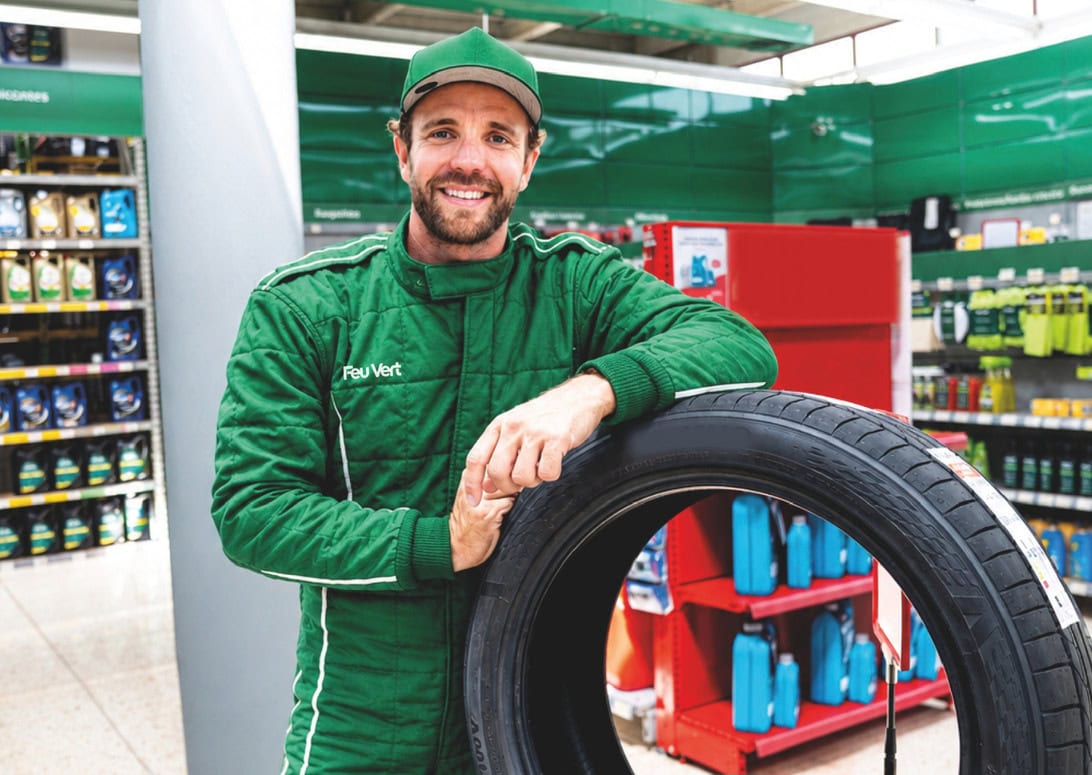 Green-uniformed mechanic smiles while resting his arm on a car tire in a hardware/auto parts store.