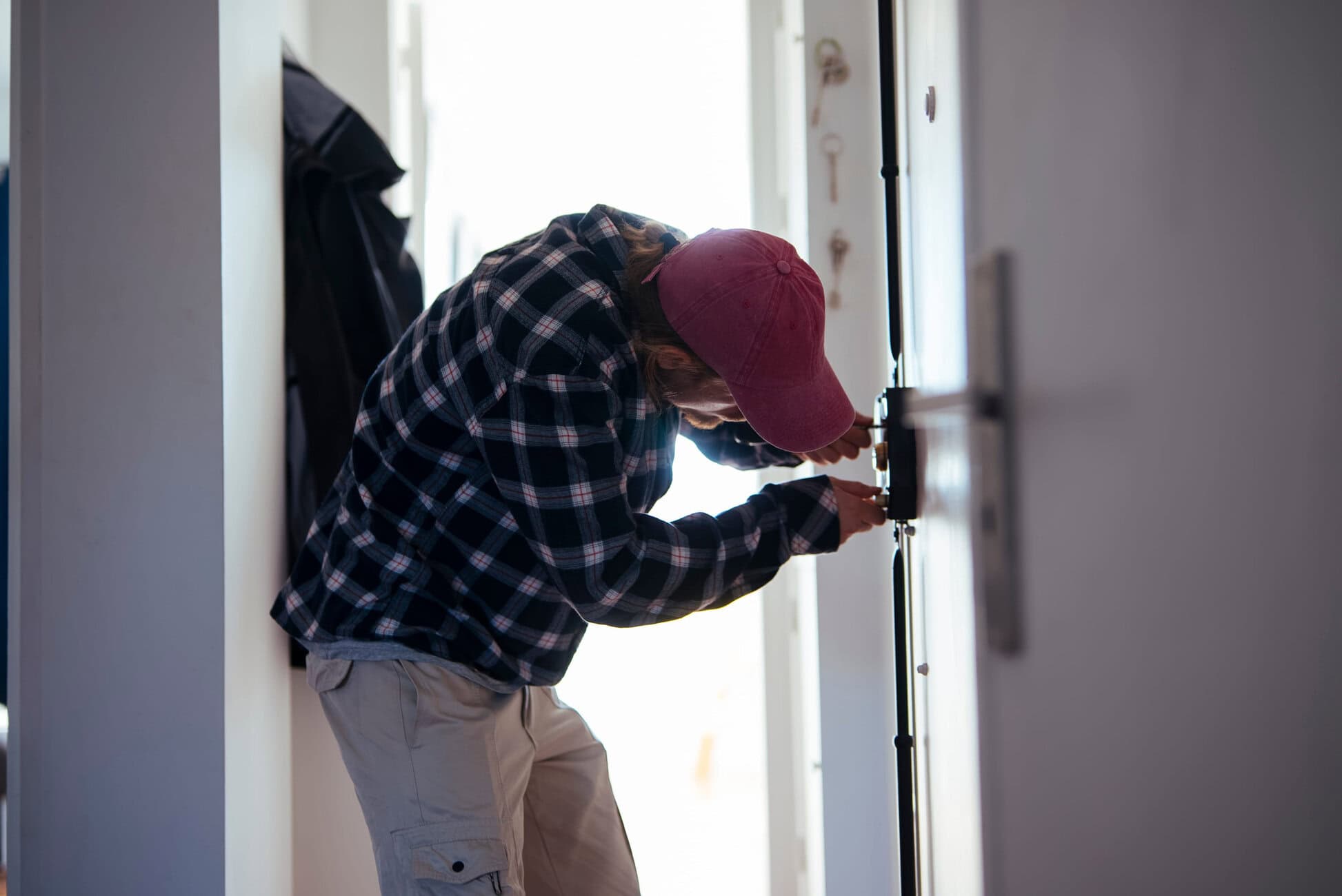 Person wearing a pink cap and plaid shirt installing or repairing a door lock in a hallway.