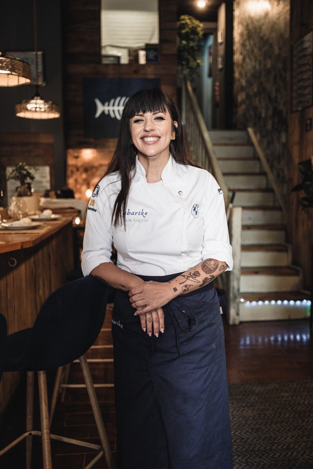 Female chef in a white jacket and navy apron, smiling at the camera in a warmly lit restaurant interior with a bar area beside her.