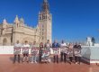 Group of people posing with posters in front of a historic stone building with a tall tower and blue sky above.