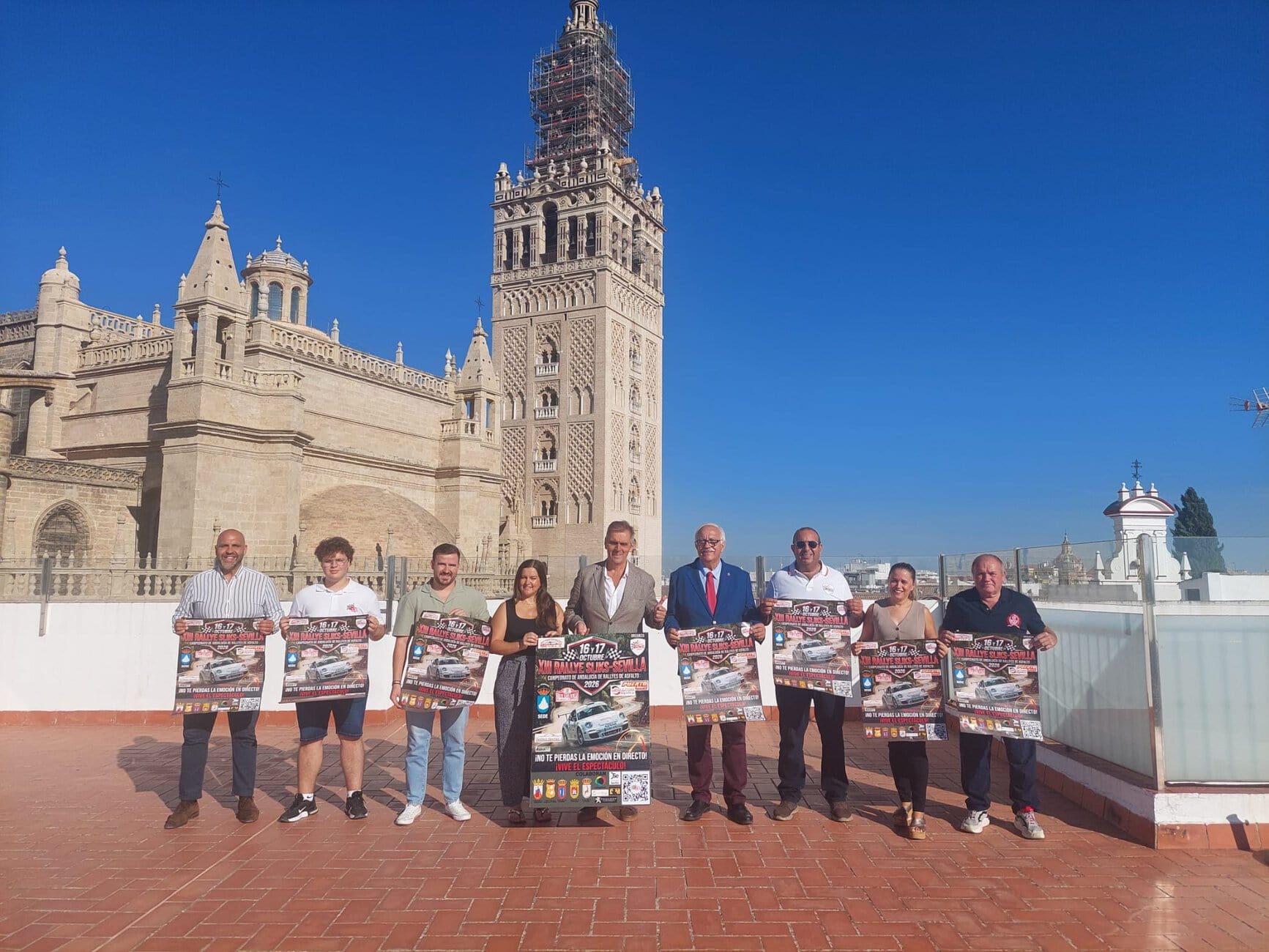 Group of people posing with posters in front of a historic stone building with a tall tower and blue sky above.