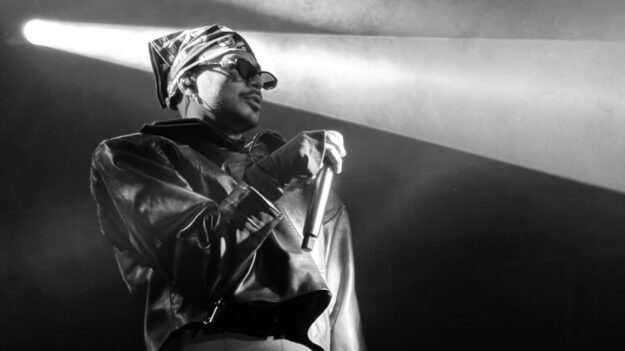 Male performer wearing sunglasses and a headwrap, holding a microphone on stage with a dramatic spotlight behind him.