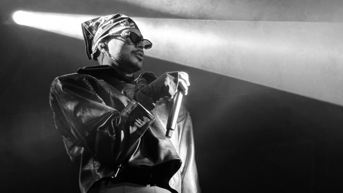Male performer wearing sunglasses and a headwrap, holding a microphone on stage with a dramatic spotlight behind him.