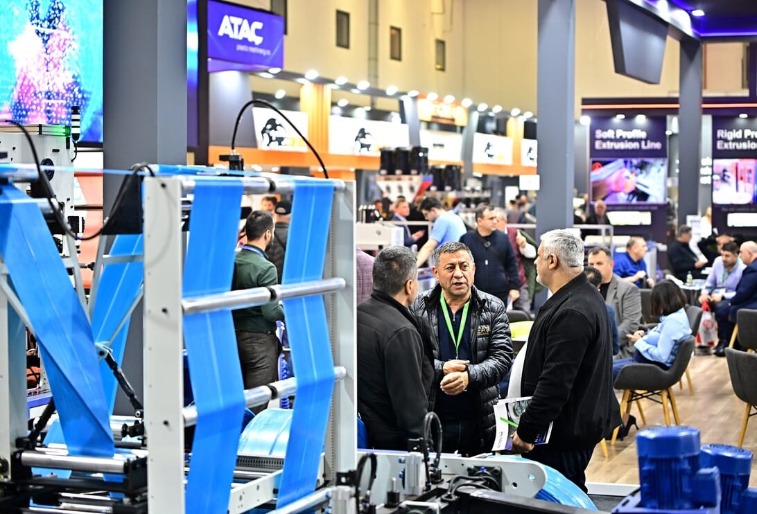 Group of men conversing near a large blue industrial machine on a trade show floor; booths and attendees visible in the background.