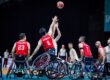 Wheelchair basketball players in red jerseys jump to reach the airborne ball during a game.