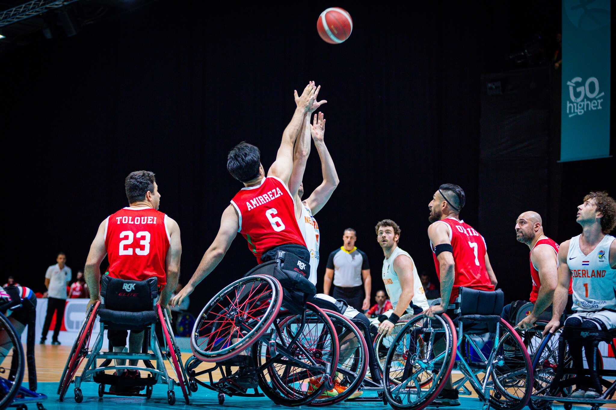 Wheelchair basketball players in red jerseys jump to reach the airborne ball during a game.