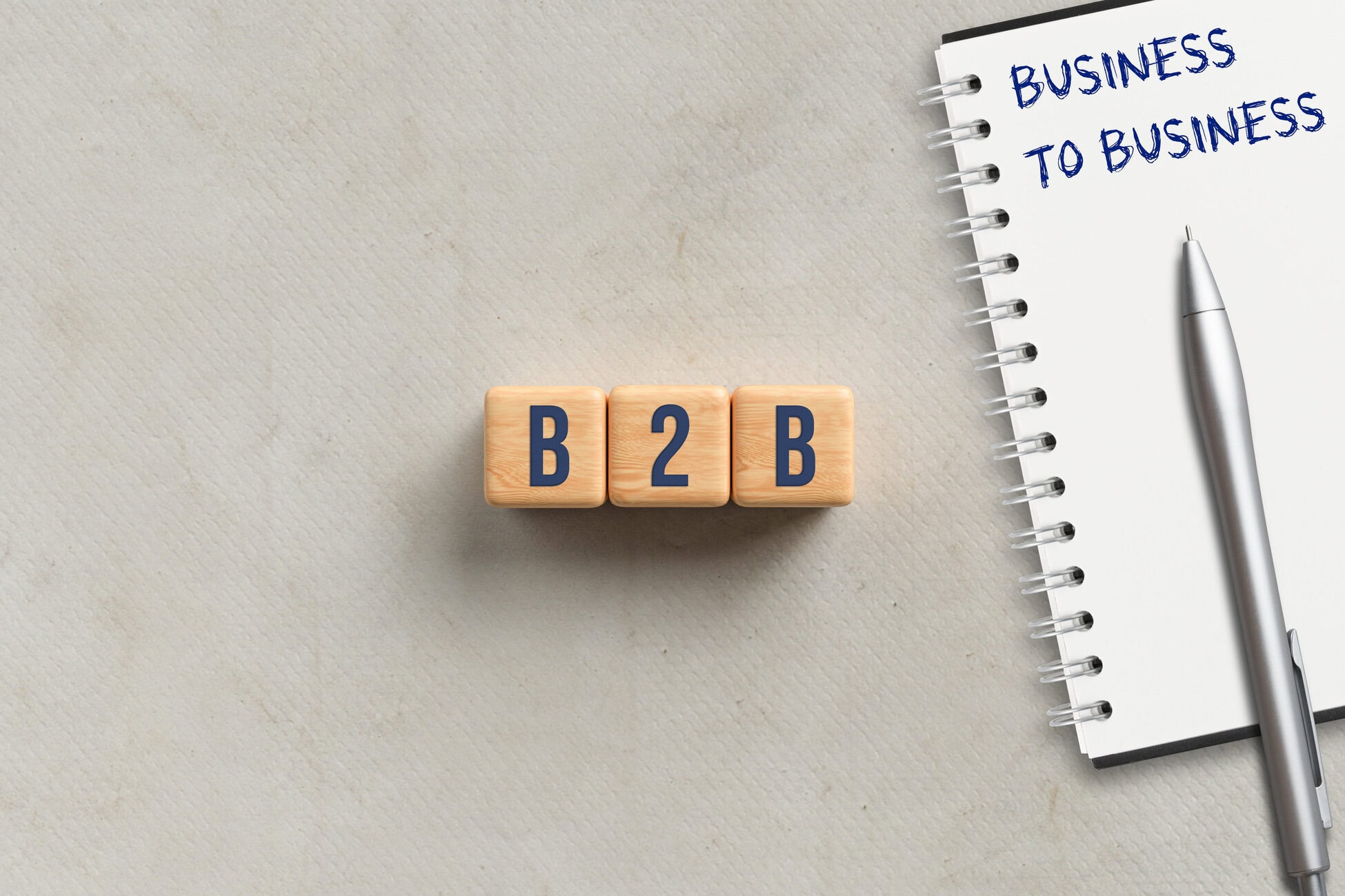 Wooden blocks spell 'B2B' beside a notebook page reading 'BUSINESS TO BUSINESS' with a silver pen nearby.