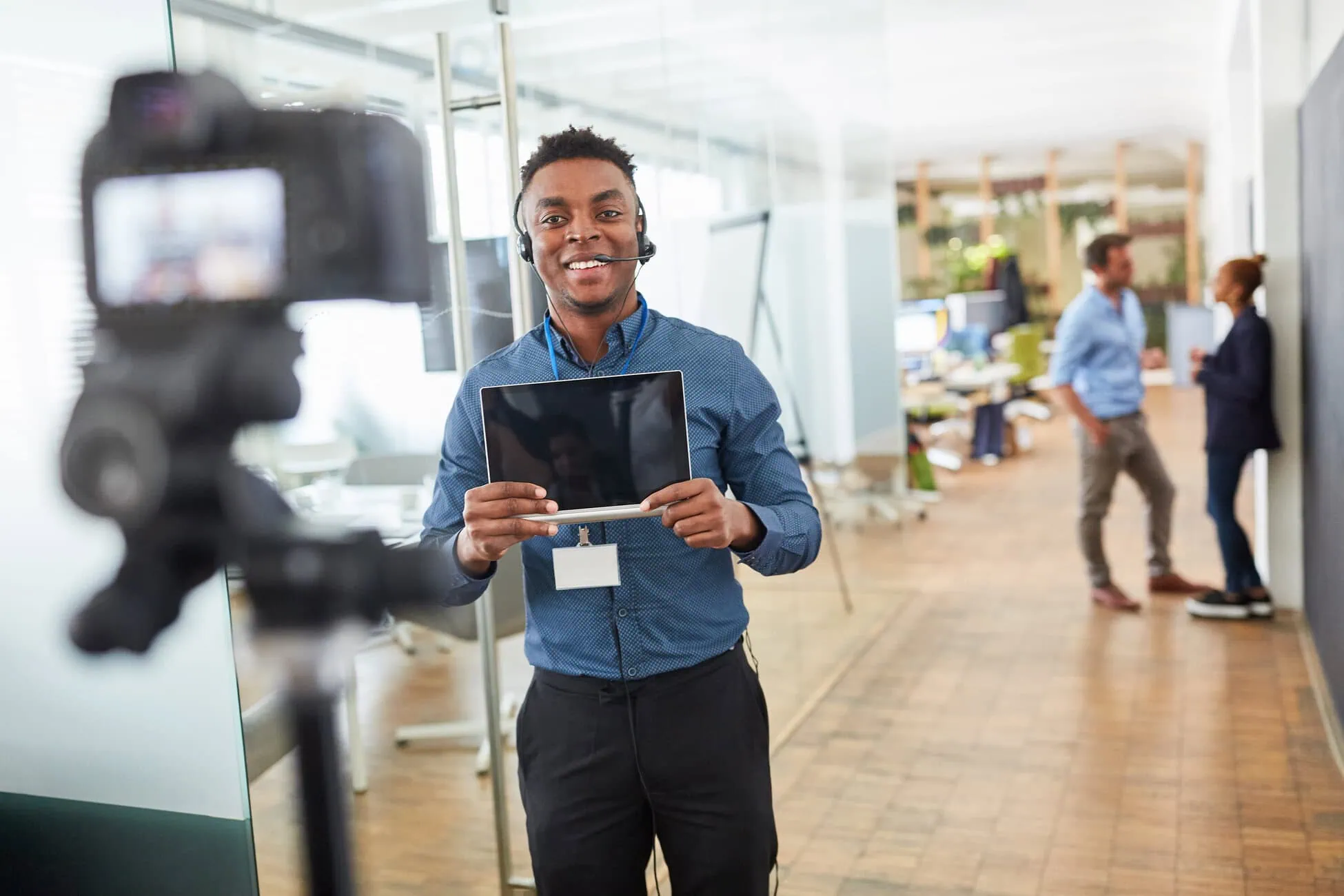 Man wearing a headset and blue shirt holds a tablet while being filmed in a bright office, with a camera in the foreground and coworkers chatting in the background.