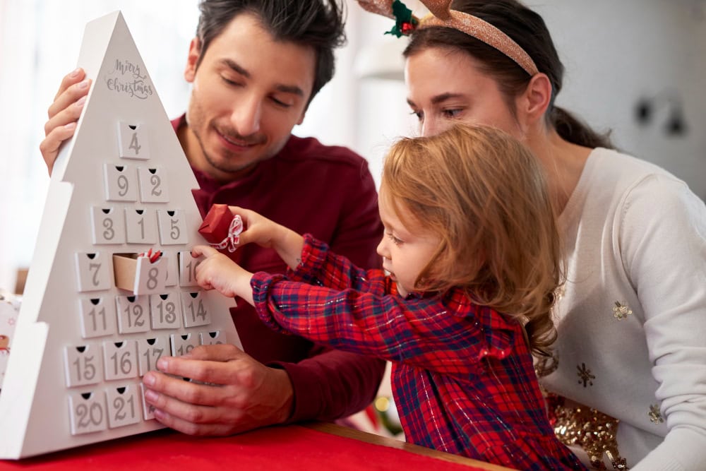 Family of three opening an Advent calendar together on a festive table, child reaching to open a door with parents nearby; holiday scene in a bright room.