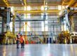Two workers in orange safety vests and blue hard hats review plans beside large yellow gantry cranes in a spacious industrial warehouse.