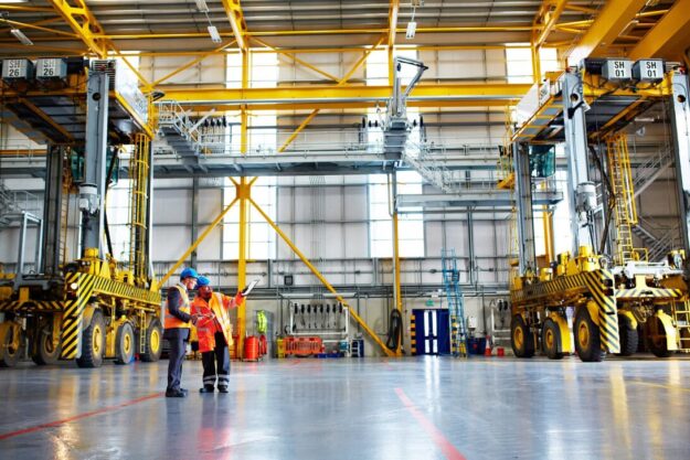 Two workers in orange safety vests and blue hard hats review plans beside large yellow gantry cranes in a spacious industrial warehouse.