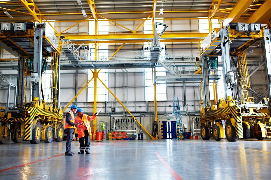 Two workers in orange safety vests and blue hard hats review plans beside large yellow gantry cranes in a spacious industrial warehouse.