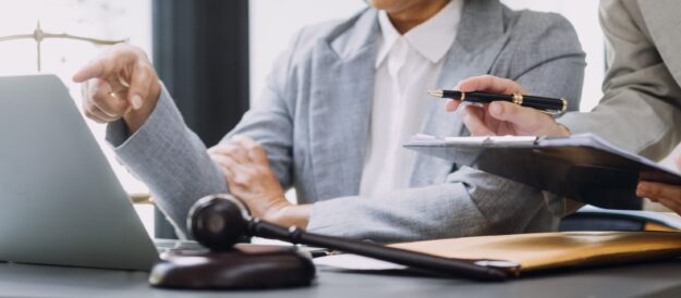 Two professionals in business attire sit at a desk with a laptop; one points toward the screen while the other holds a pen and clipboard, discussing documents.
