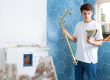 Man in a white t-shirt installs a brass showerhead on a blue hex-tile wall outdoors/renovation scene.