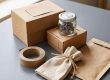 Packing scene with stacked cardboard boxes, a glass jar of seeds, and nearby packing supplies on a grey surface.