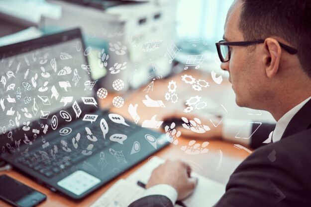 Businessman in a suit typing on a tablet at a desk with white digital icons overlaying the scene