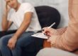 Therapist writing on a clipboard during a therapy session, while a man sits in the background with his hand on his head.