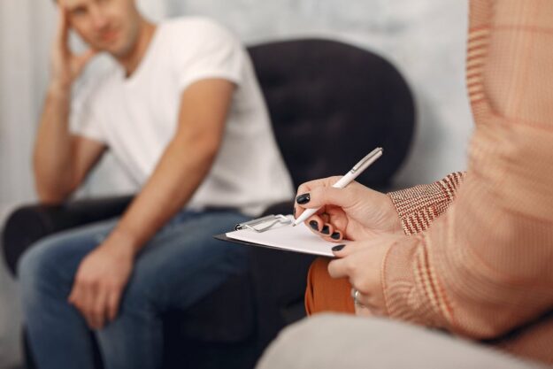 Therapist writing on a clipboard during a therapy session, while a man sits in the background with his hand on his head.