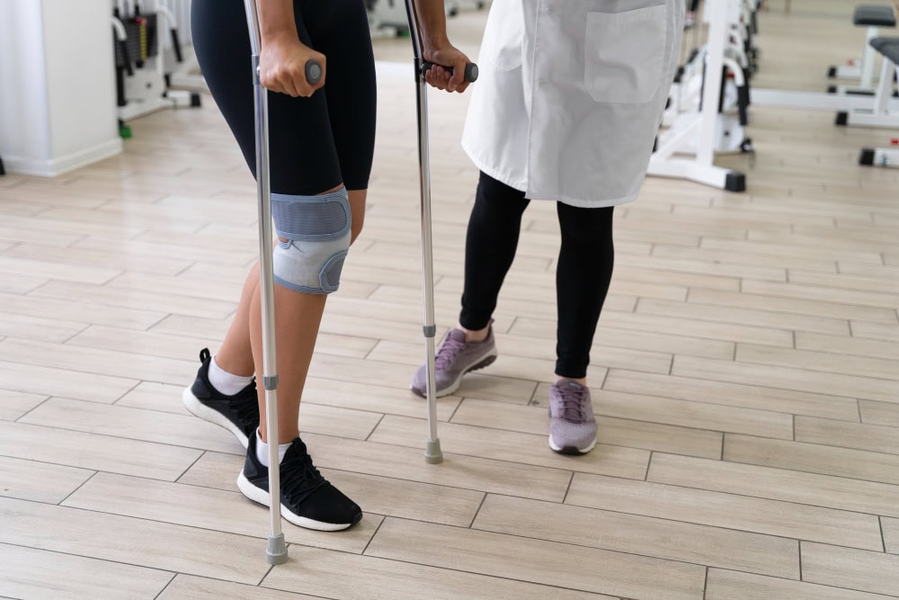 Person with a knee brace using crutches during physical therapy, with a clinician nearby for support.