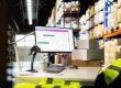 Warehouse desk with computer showing package tracking, surrounded by stacked boxes and tall shelves in the background.