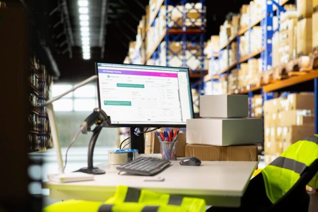 Warehouse desk with computer showing package tracking, surrounded by stacked boxes and tall shelves in the background.