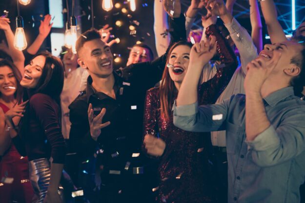 Group of friends cheering and laughing at a lively party with confetti and warm hanging lights in the foreground and background.