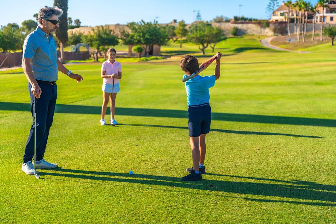 Adult male golf instructor or parent watches a boy in a blue shirt swing a club on a sunny greens golf course while a girl in pink stands nearby observing.