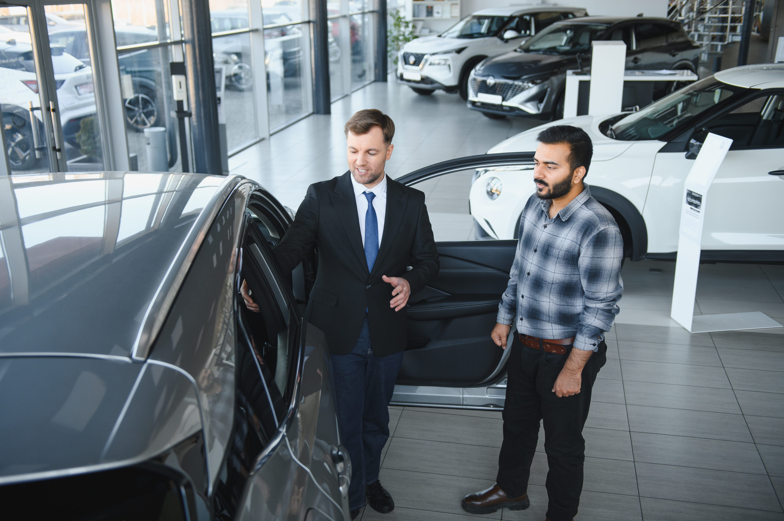 Two men in a car showroom with one opening a car door to inspect the interior while others look on.