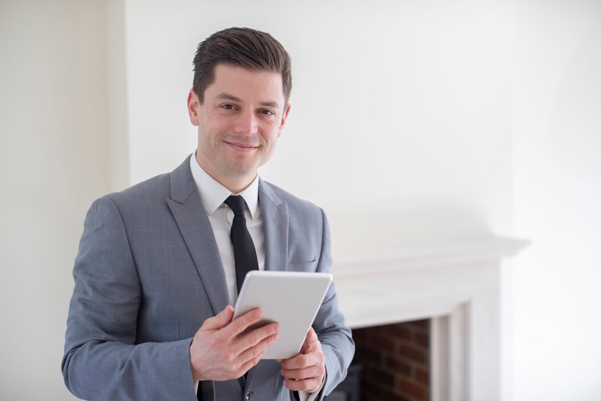 Smiling businessman in a gray suit and black tie holds a tablet indoors by a white wall.