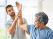 Older man with gray hair performs an arm raise guided by a younger therapist in a bright clinic room.