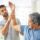 Older man with gray hair performs an arm raise guided by a younger therapist in a bright clinic room.