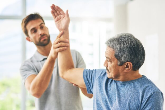 Older man with gray hair performs an arm raise guided by a younger therapist in a bright clinic room.