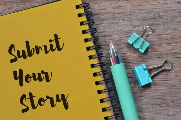 Yellow spiral notebook reading 'Submit Your Story' beside a teal pen and binder clips on a wooden desk.