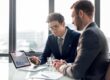 Two businessmen in suits discuss a tablet showing a graph during a meeting in a bright office.