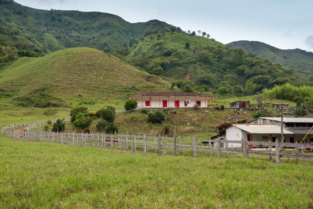 Green rolling hills and meadows with a small white building featuring red doors, bordered by a winding wooden fence and farm structures nearby.