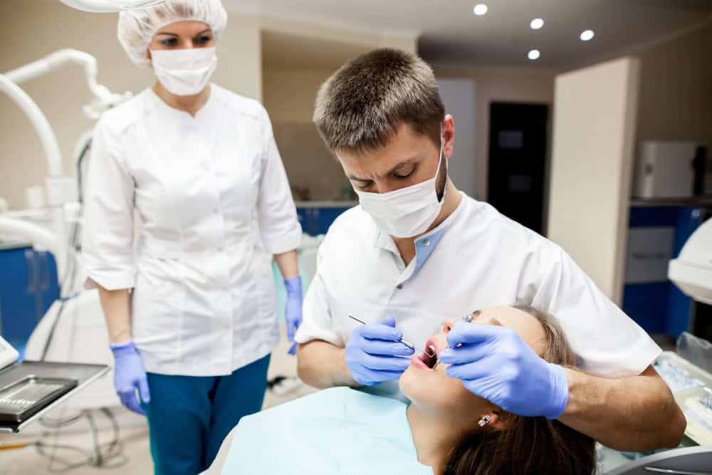 Dentist wearing a mask and gloves treats a patient, using dental tools while an assistant stands nearby in a clinical office.