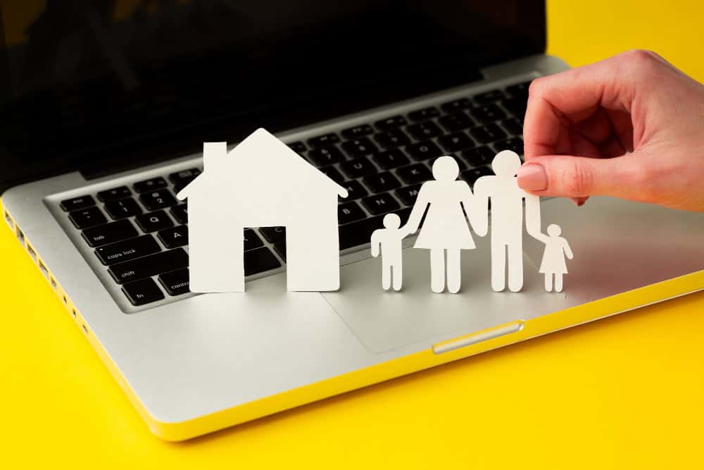 Paper cutouts of a house and a family being held by a hand on a laptop keyboard, on a bright yellow background — symbolizing online home/family protection.