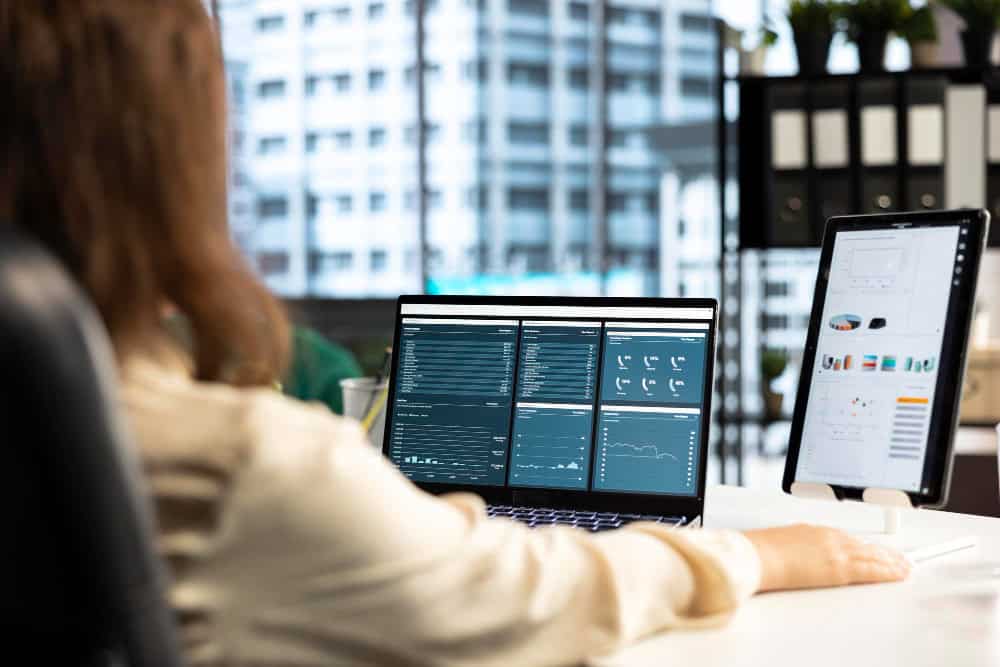 Person sits at a desk with a laptop showing analytics dashboards and a tablet displaying charts in a modern office.