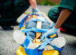 Paramedics secure an injured person on a yellow stretcher with blue straps on a street side.