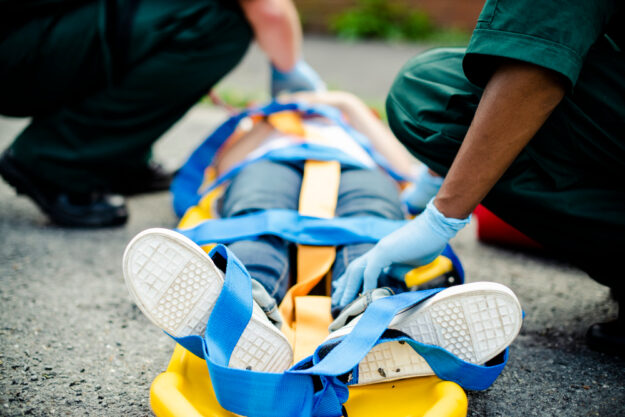 Paramedics secure an injured person on a yellow stretcher with blue straps on a street side.