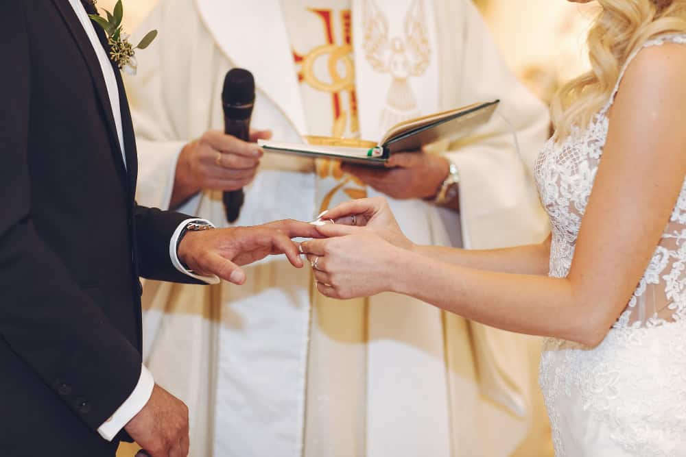 Bride and groom exchange rings during a wedding ceremony, with a clergy member holding a book in the background