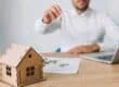 Man in a white shirt holds up a key over a miniature wooden house on a desk with documents and a laptop nearby, suggesting a real estate transaction.