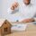 Man in a white shirt holds up a key over a miniature wooden house on a desk with documents and a laptop nearby, suggesting a real estate transaction.