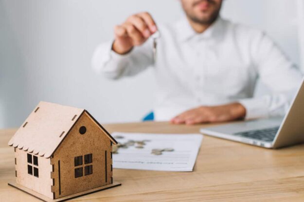 Man in a white shirt holds up a key over a miniature wooden house on a desk with documents and a laptop nearby, suggesting a real estate transaction.