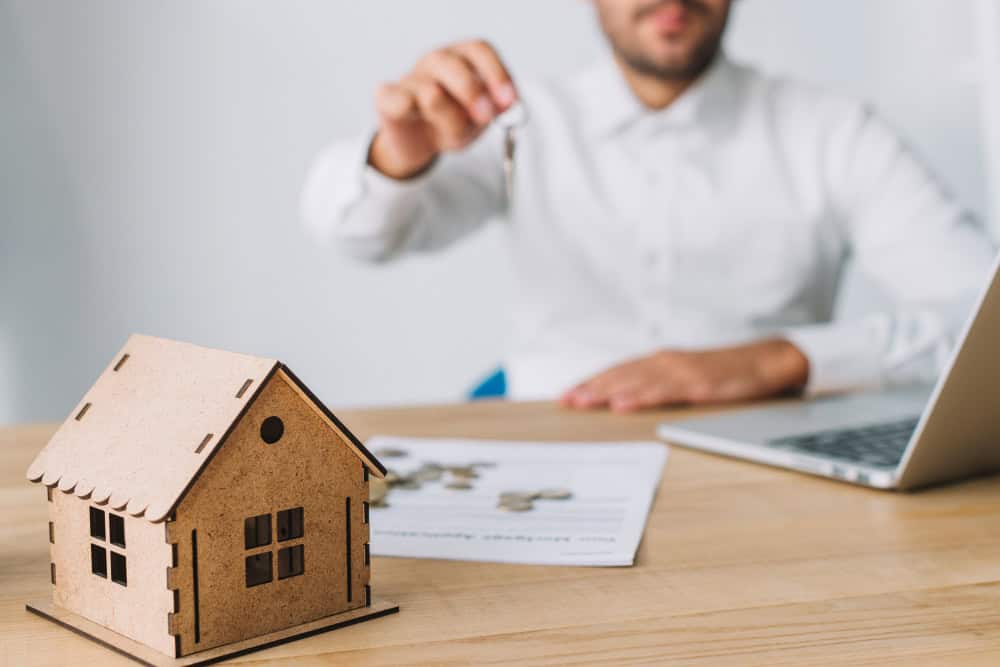 Man in a white shirt holds up a key over a miniature wooden house on a desk with documents and a laptop nearby, suggesting a real estate transaction.