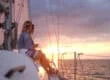 Two people sit on the edge of a sailboat at sunset, sharing a drink while gazing toward the horizon over calm water.