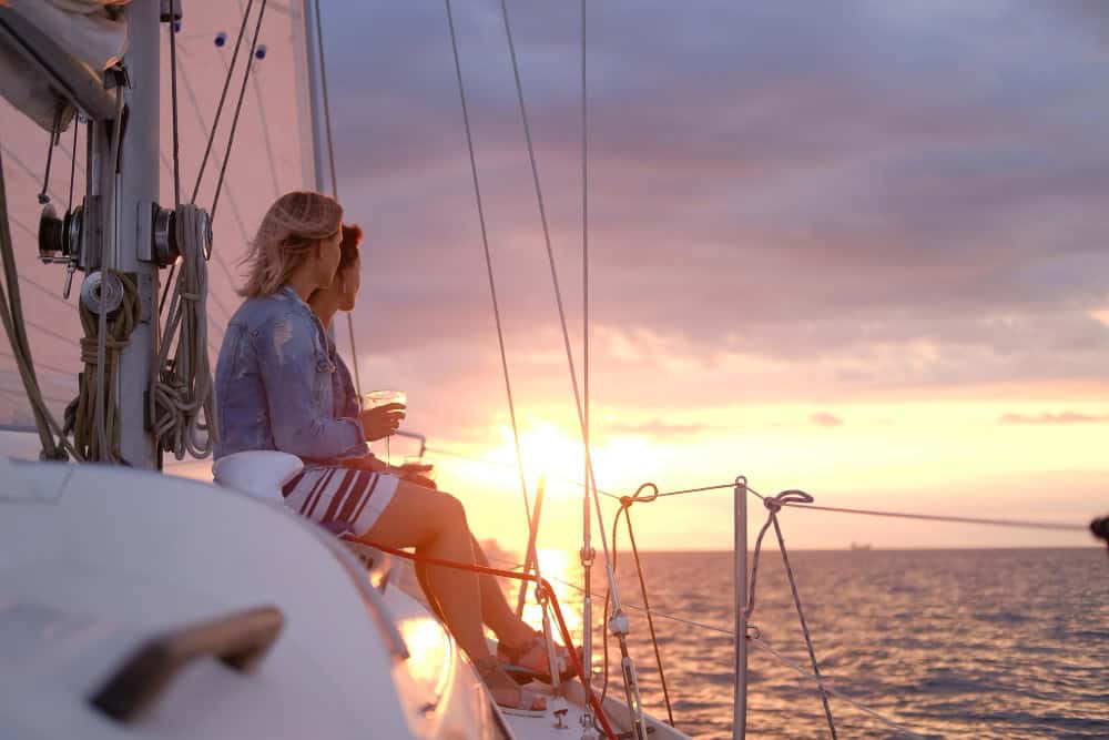 Two people sit on the edge of a sailboat at sunset, sharing a drink while gazing toward the horizon over calm water.