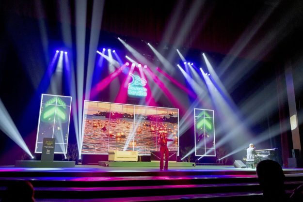 Stage with colorful blue, purple, and white spotlights; large screens show a sunset over water while a singer performs on stage.