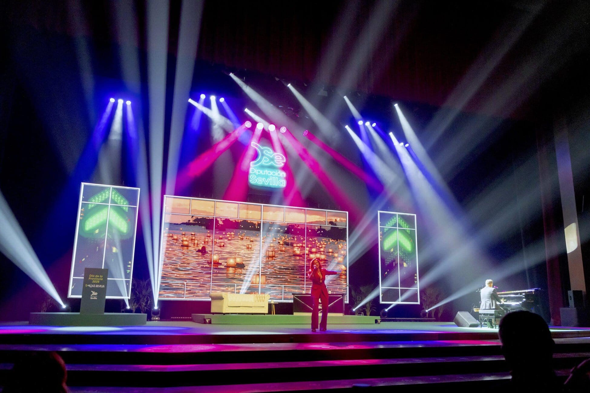 Stage with colorful blue, purple, and white spotlights; large screens show a sunset over water while a singer performs on stage.