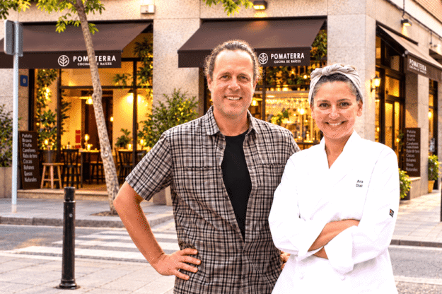 Chef Ana Diaz and a man in a plaid shirt pose outside Pomaterra restaurant on a sunny street.